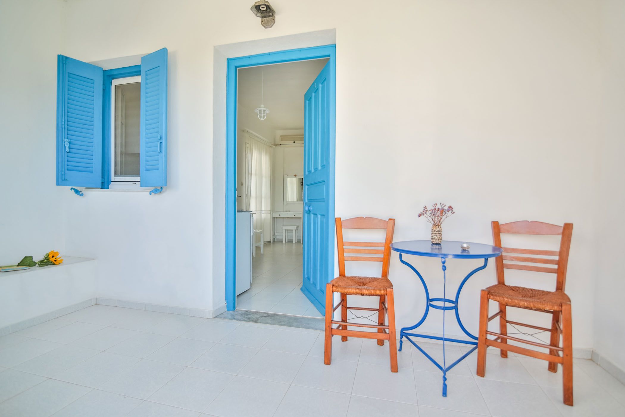Una habitación con piso de baldosas blancas, una puerta azul y una mesa azul con dos sillas.