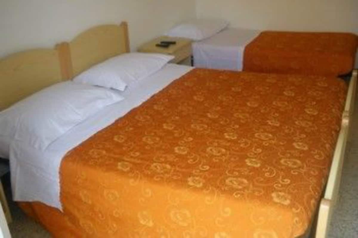 A single bed with an orange bedspread and white pillows in a room with a plain wall and a small nightstand.