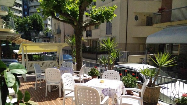 An outdoor patio with white plastic chairs and tables, surrounded by potted plants and a large tree. - Hotel Ronconi (Rimini)