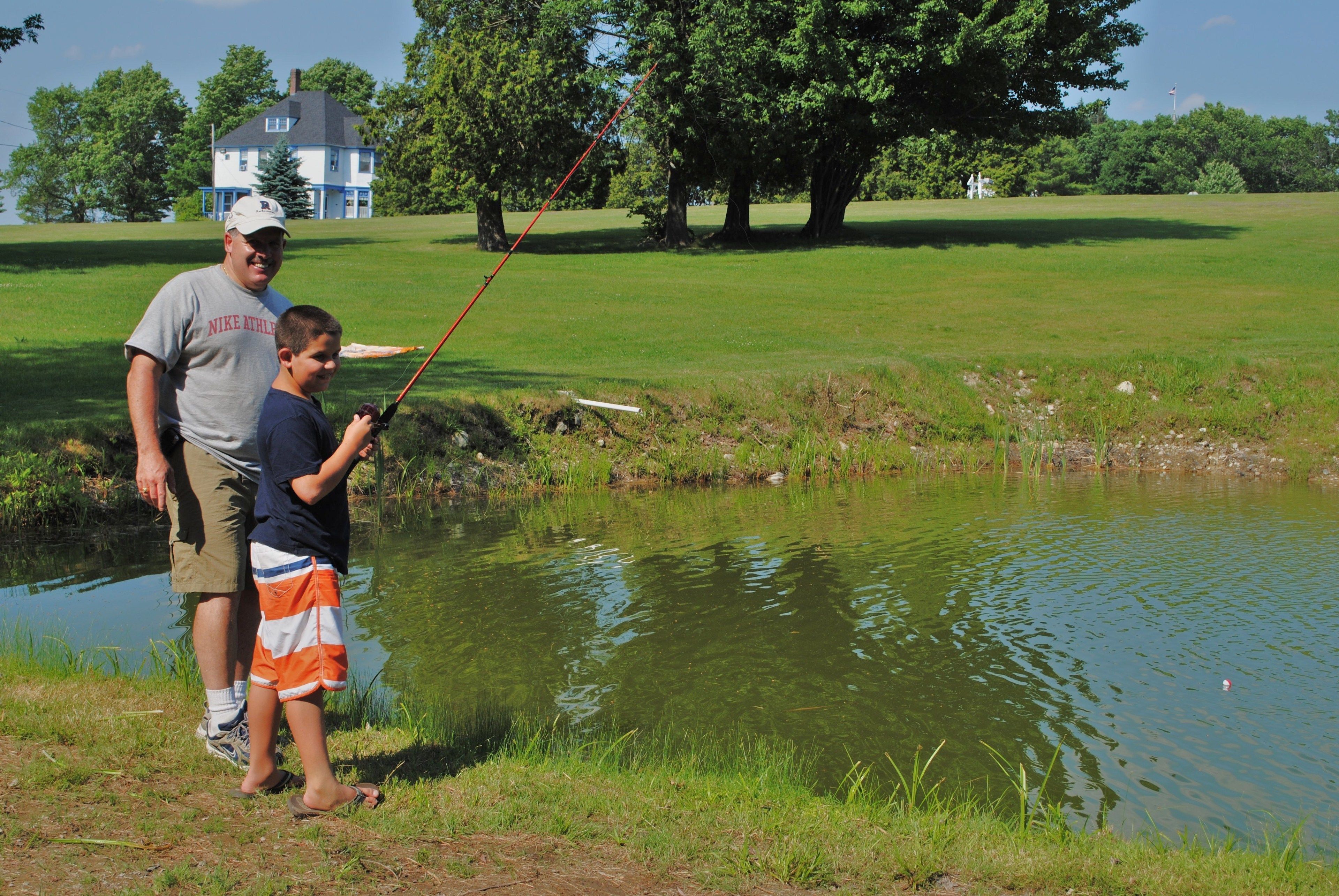 A man and a boy fishing by a pond with a house in the background.