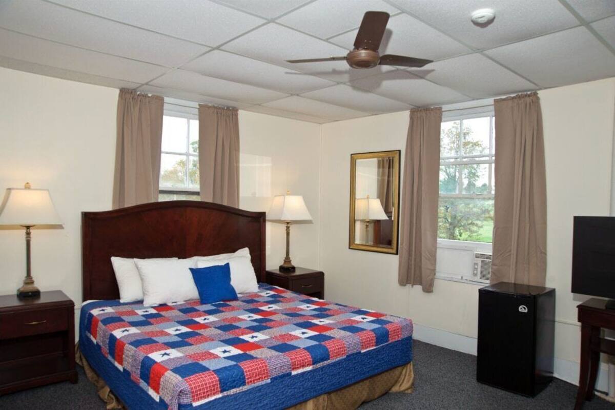 A hotel room with a wooden bed, two bedside lamps, a ceiling fan, a TV, and a window with curtains.