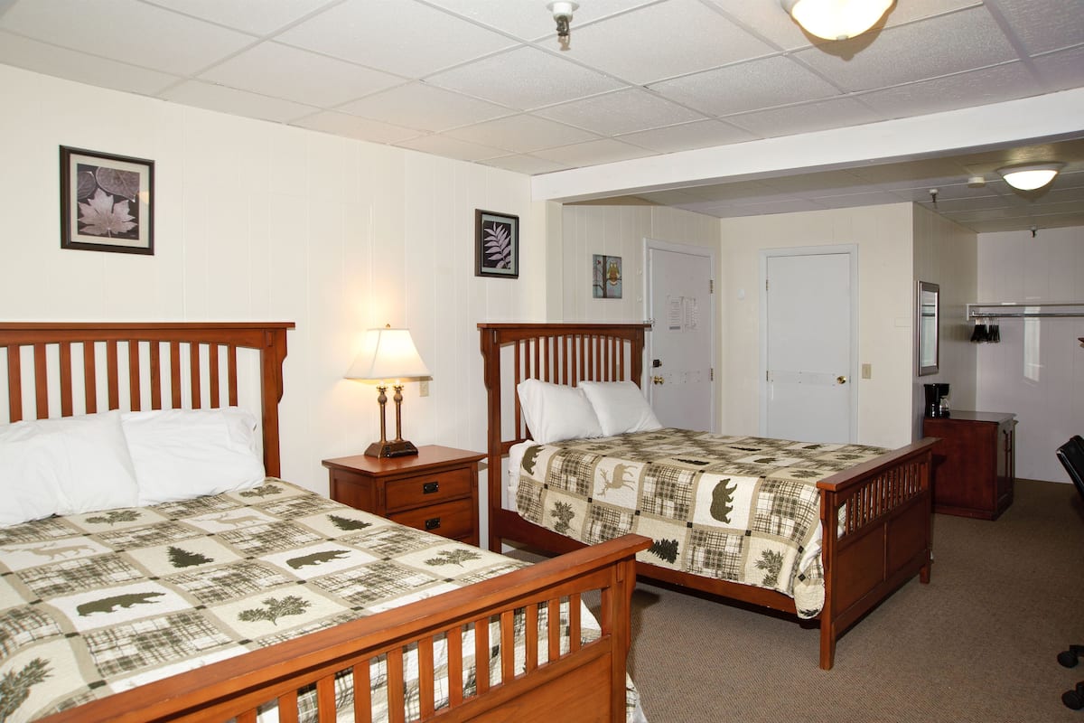 A hotel room with two wooden beds, a nightstand with a lamp, a dresser, and a door leading to a bathroom.