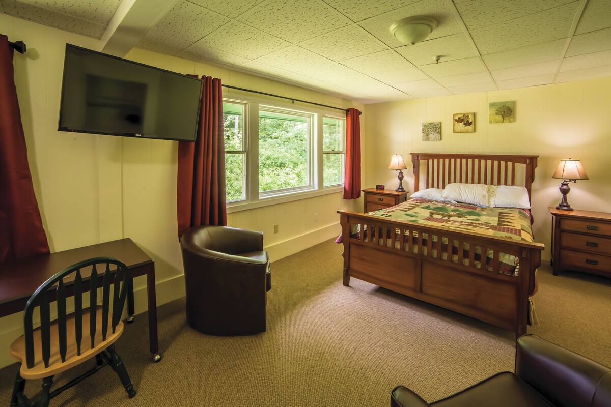 A bedroom with a bed, a desk, a chair, a TV mounted on the wall, and two windows with curtains.