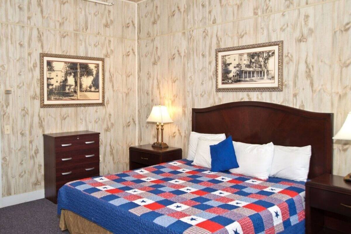 A bedroom with a wooden headboard, a quilted bedspread, a nightstand with a lamp, and two framed pictures on the wall.