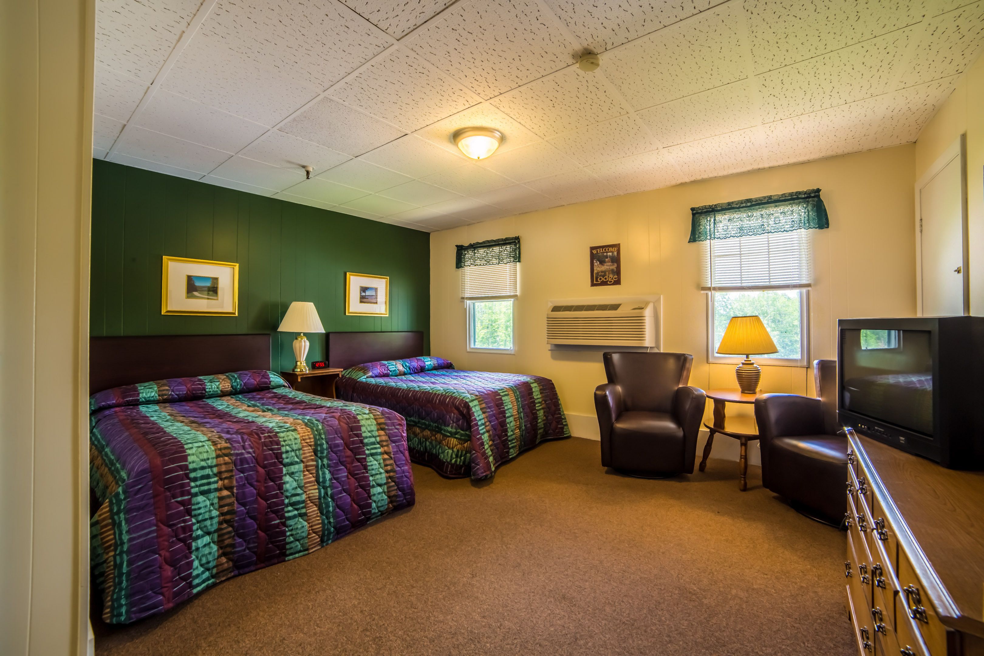 A room with two beds, a television, a chair, and a table with a lamp.