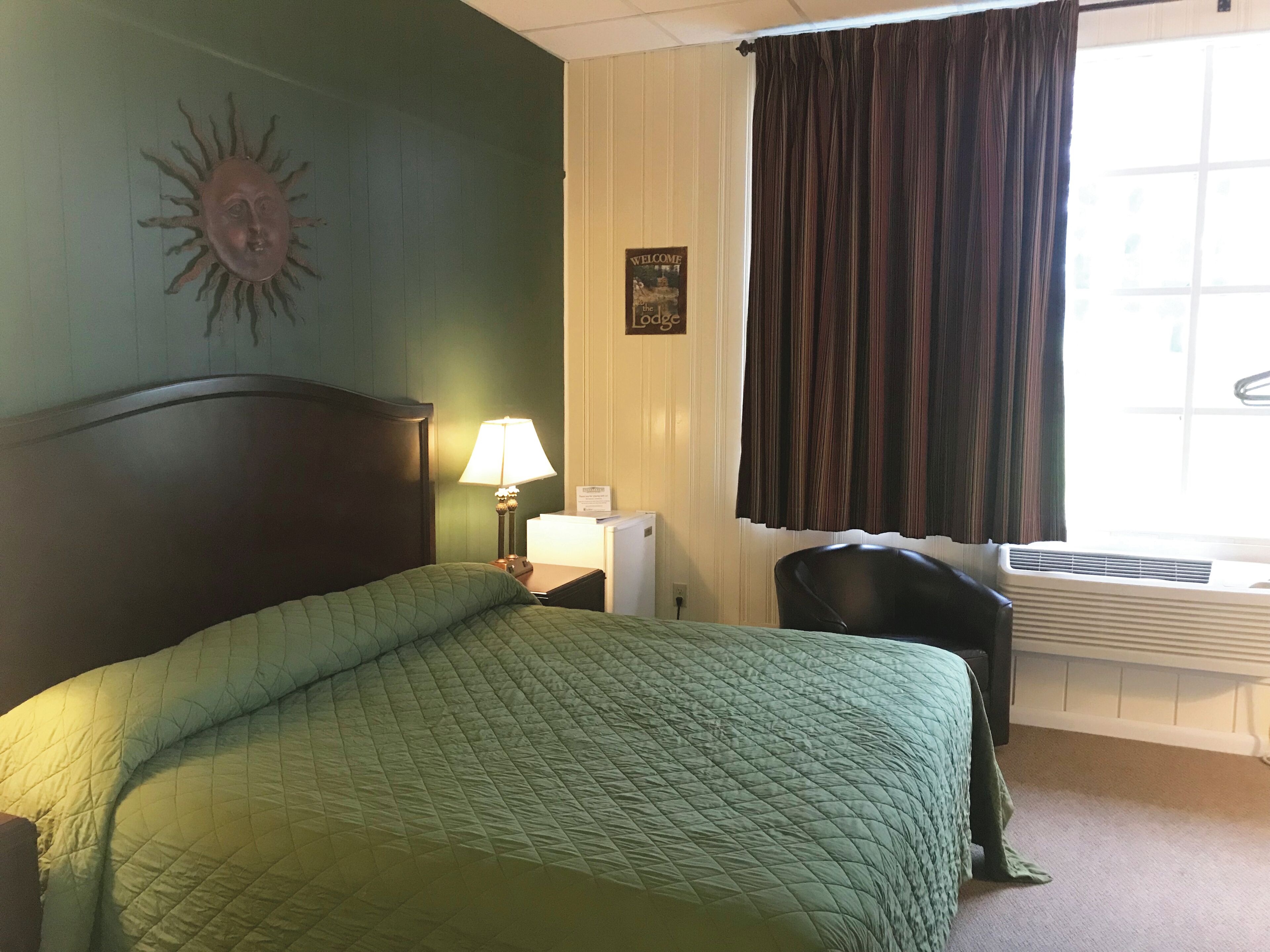 A hotel room with a bed, a nightstand, a lamp, a chair, a window with curtains, and a wall decoration.