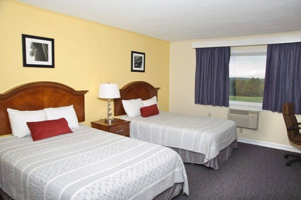 A hotel room with two beds, a desk with a lamp, a chair, and a window with curtains.