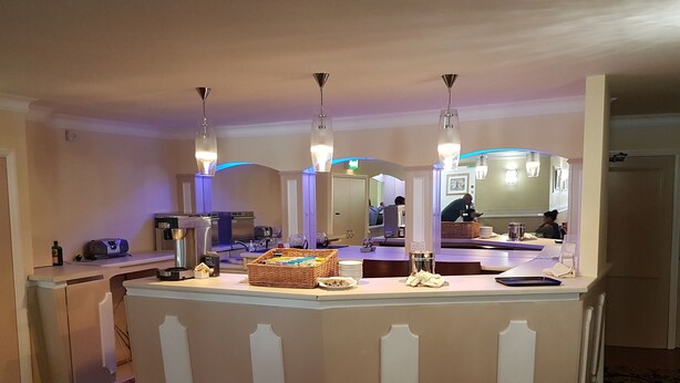 A hotel bar area with a central counter, multiple light fixtures, and a large mirror. - Maison Gorey Hotel (Grouville)