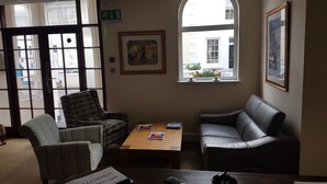 A waiting area with a sofa, armchair, and a small table with magazines. There is a window with a view of buildings outside. - Maison Gorey Hotel (Grouville)