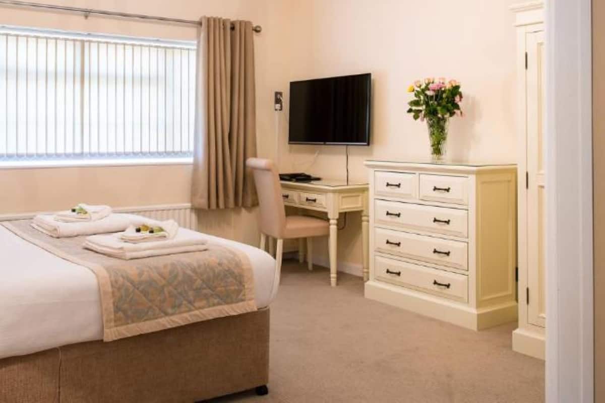 Ein Hotelzimmer mit Bett, Nachttisch, Lampe, Schreibtisch, Stuhl, Fernseher, Kommode und einem Fenster mit Vorhängen.