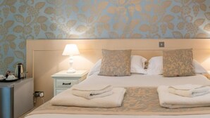 A hotel room with a large bed, two bedside lamps, a nightstand with a coffee maker, and a floral wallpaper. - Maison Gorey Hotel (Grouville)