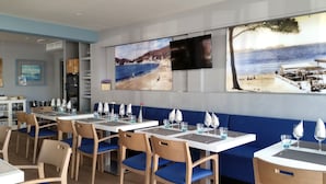 A restaurant with blue cushioned booths, white tables, and chairs. There are paintings of coastal scenes on the walls. - Hotel Creta Paguera (Calvia)