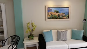 A living room with a white sofa, a small table with a potted plant, a black chair, and a painting of a coastal village on the wall. - Hotel Creta Paguera (Calvia)