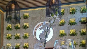 Ένα εσωτερικό εστιατορίου με ένα ξύλινο τραπέζι, δύο ποτήρια κρασιού, ένα κεντρικό φυτό ορχιδέας και έναν τοίχο από πέτρα με πράσινα φυτά.
