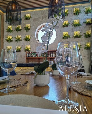 A restaurant interior with a wooden table, two wine glasses, a central orchid plant, and a stone wall with green plants. - Alesia Hotel Boutique & Spa (Aguascalientes)