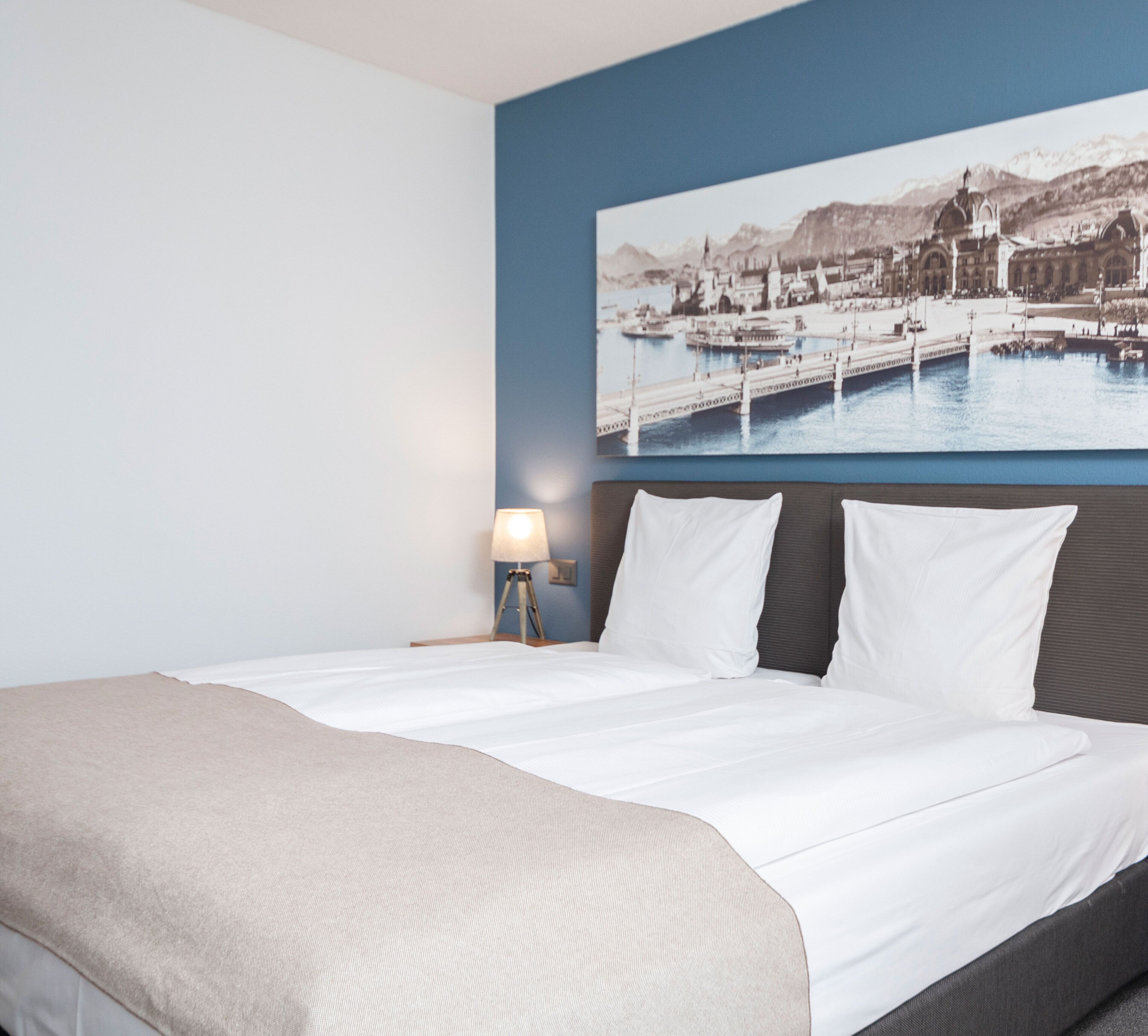 A hotel room with a bed, a nightstand, a lamp, and a wall-mounted picture of a harbor.