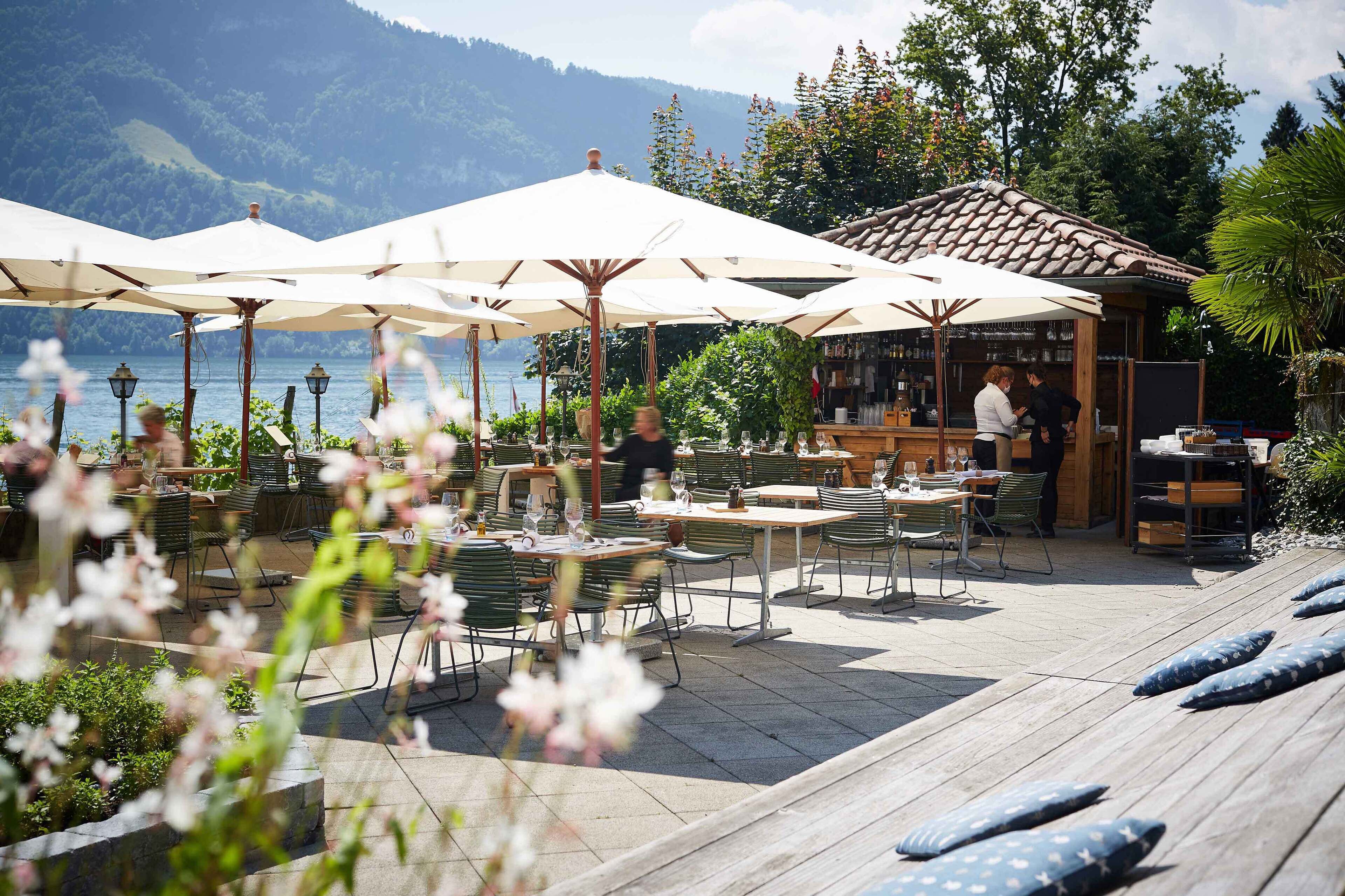 Un espacio exterior para comer con mesas y sombrillas, con vista a un lago y montañas al fondo.