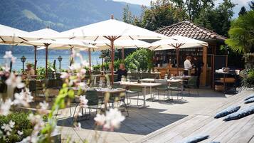Un espacio exterior para comer con mesas y sombrillas, con vista a un lago y montañas al fondo.