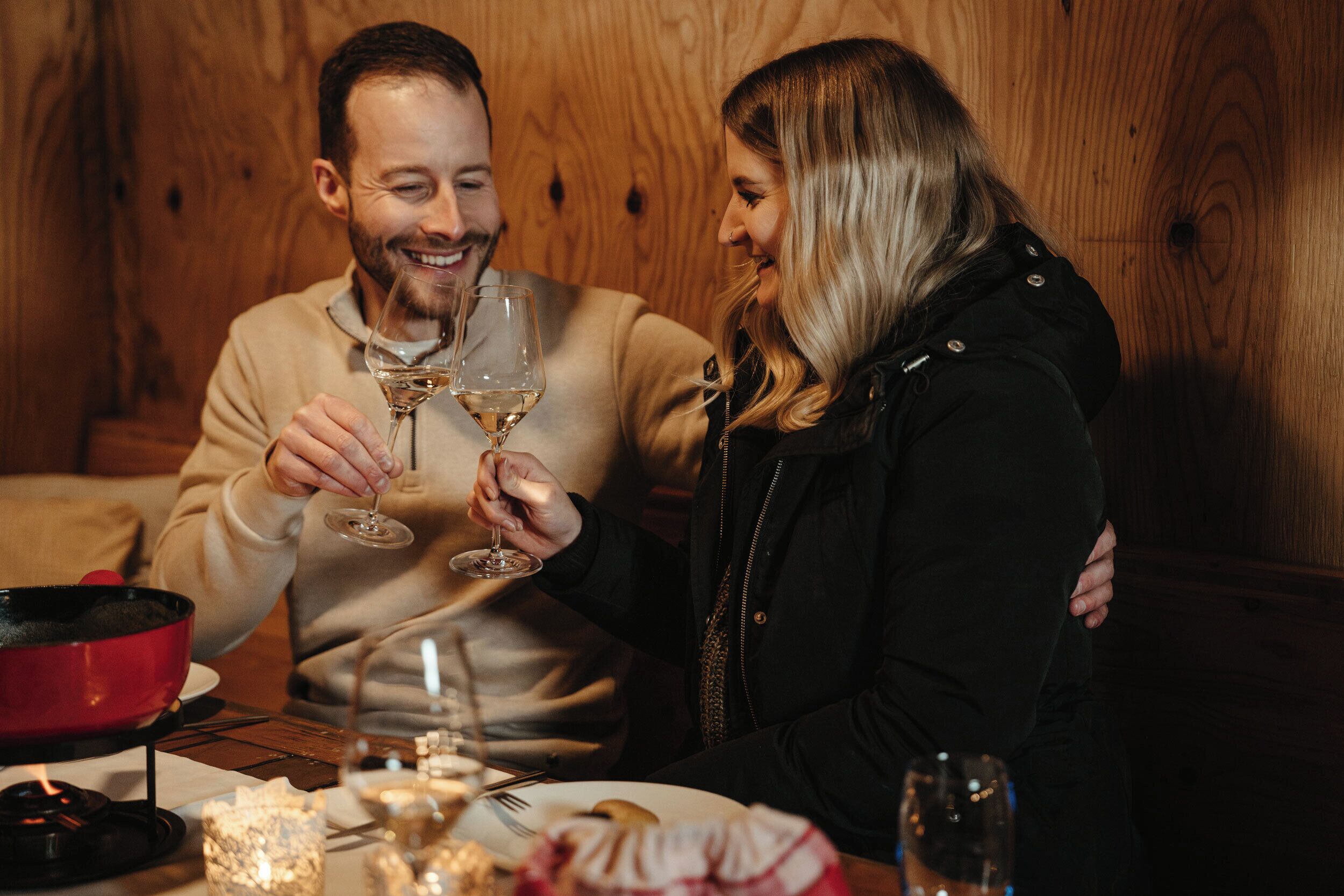 Un couple est assis à une table en bois, devant des verres à vin, un pot rouge et une bougie.