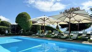 A swimming pool with lounge chairs and umbrellas, surrounded by greenery and a clear sky. - Seehotel Kastanienbaum (Horw)