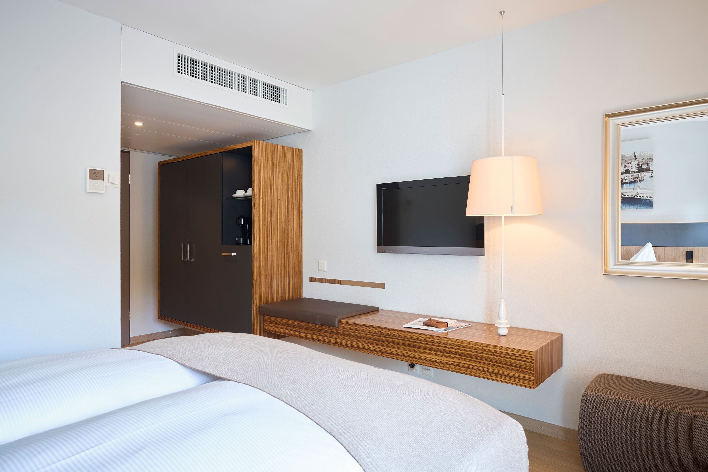 A hotel room with a bed, a TV mounted on the wall, a bedside table with a lamp, and a wardrobe.