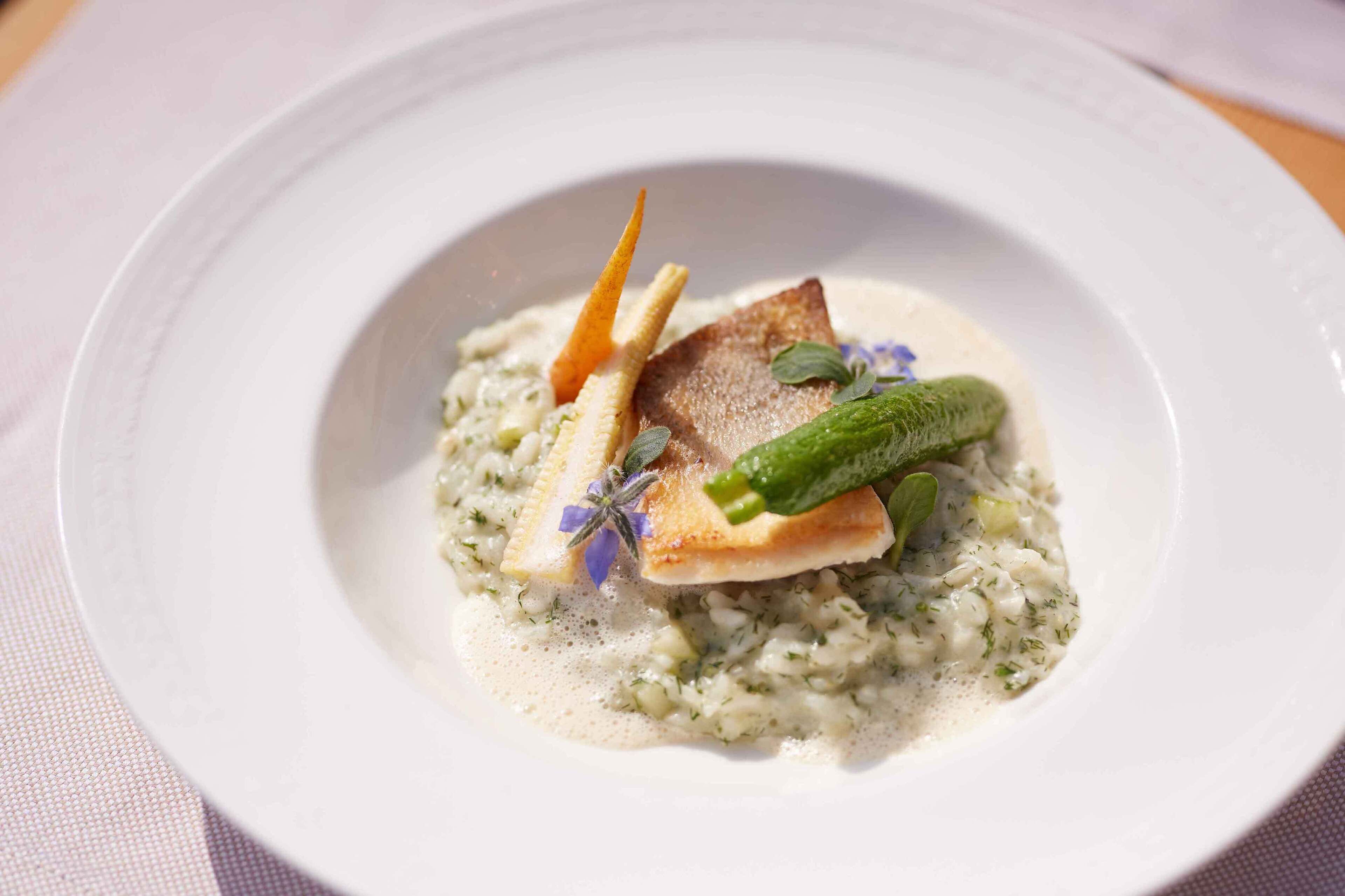 Un plato blanco con guarnición de pescado, verduras verdes y salsa cremosa, adornado con hierbas y una rebanada de pan.