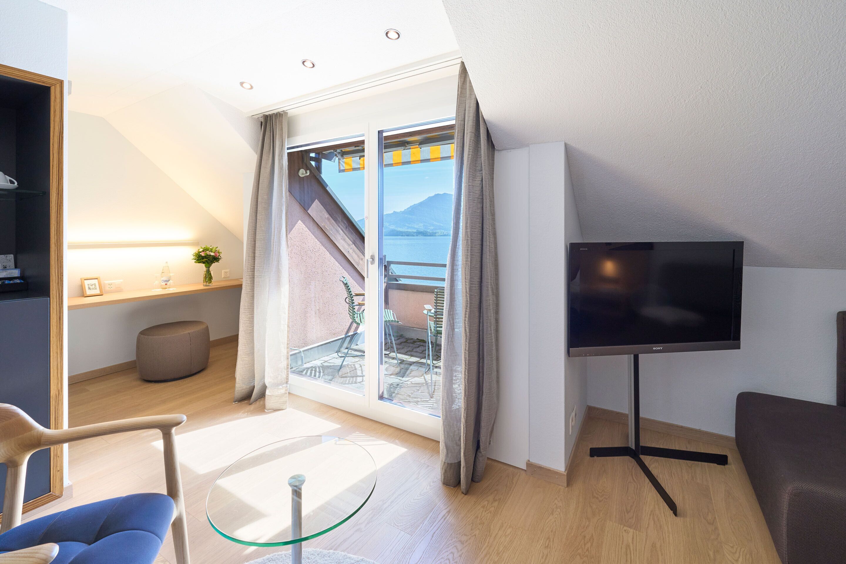 A modern room with a TV, a glass table, a sofa, a chair, a small ottoman, and a sliding glass door leading to an outdoor area with chairs.