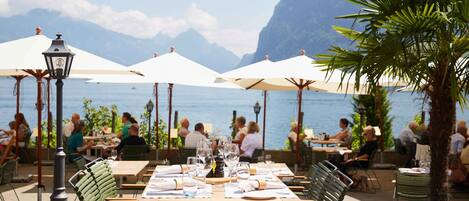 An outdoor dining area with tables and chairs, umbrellas, and a view of the sea and mountains.
