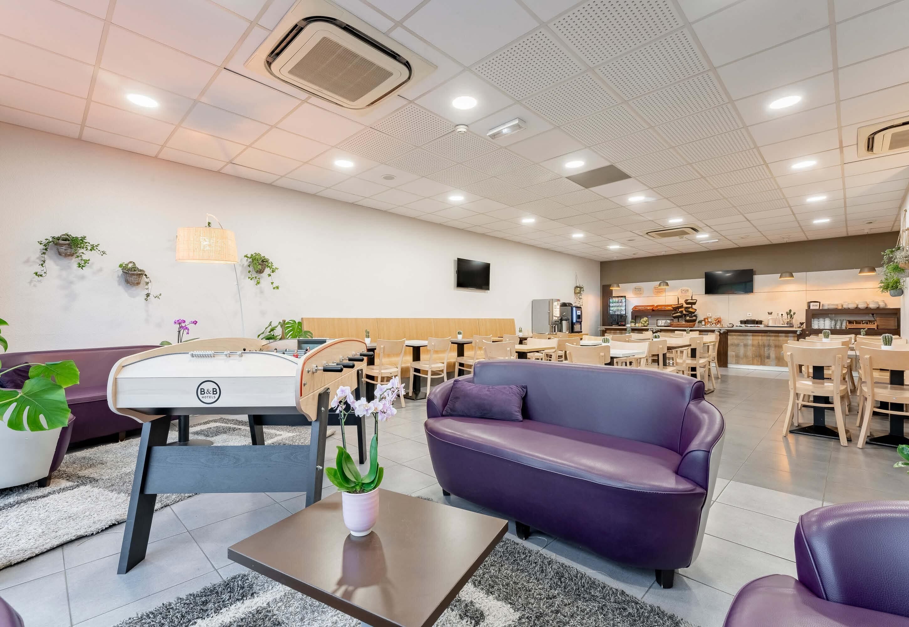 Een moderne hotellobby met een paarse bank, een houten tafel met een bloevas, en een zithoek met stoelen en tafels.