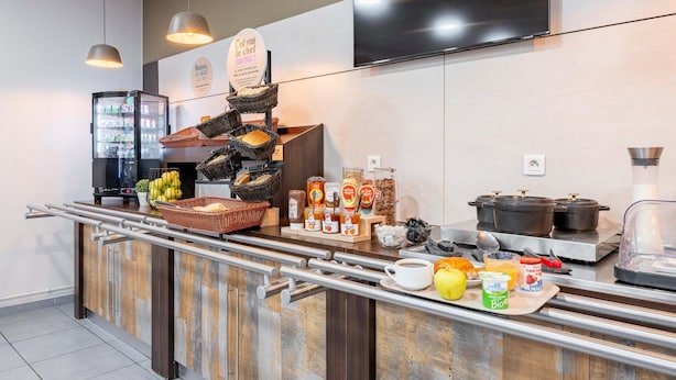 A breakfast bar with a variety of food items, a coffee station, and a TV mounted on the wall. - B&B HOTEL Maubeuge Louvroil (Louvroil)