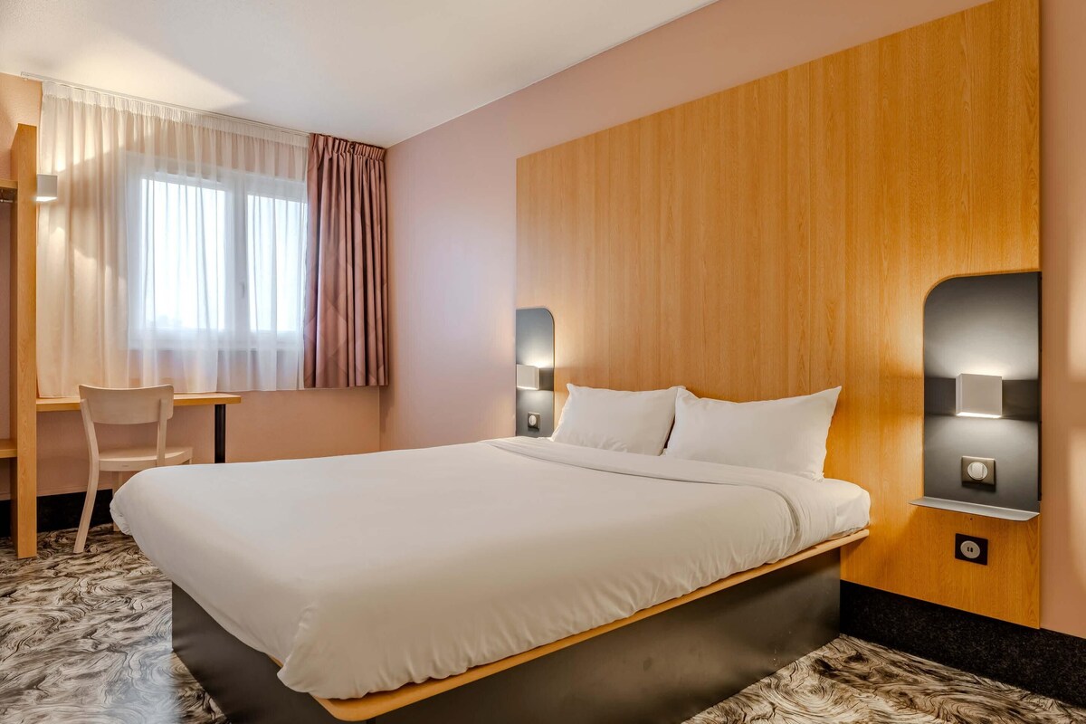 A hotel room with a large bed, a desk, a chair, and two wall-mounted lights.