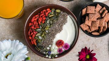 Un tazón de yogur cubierto con semillas de chía, semillas de calabaza y bayas de goji, rodeado de flores y un vaso de jugo de naranja.