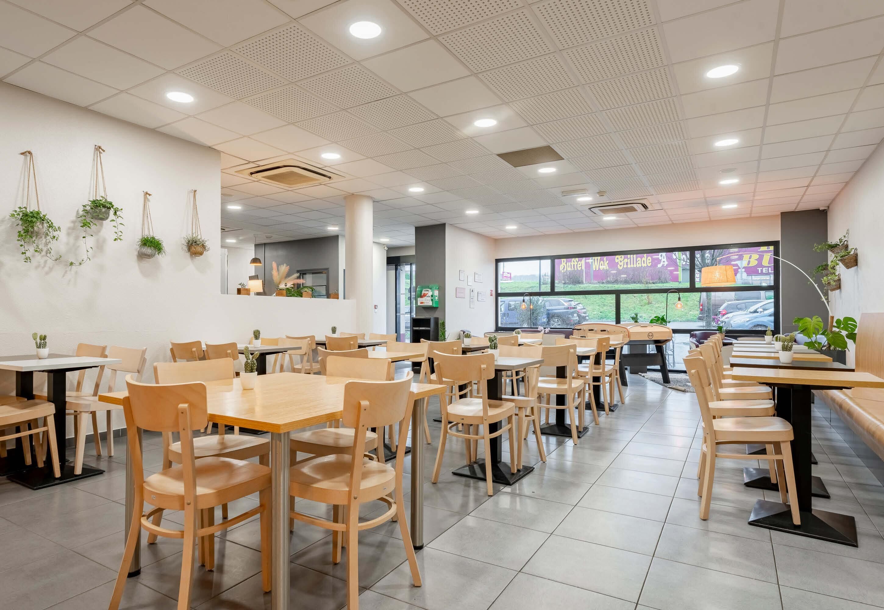 Interior de restaurante con mesas y sillas de madera, luces de techo y plantas colgantes.