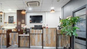 A reception area with a wooden reception desk, a flat-screen TV, a bar area with a menu, and a potted plant. - B&B HOTEL Maubeuge Louvroil (Louvroil)