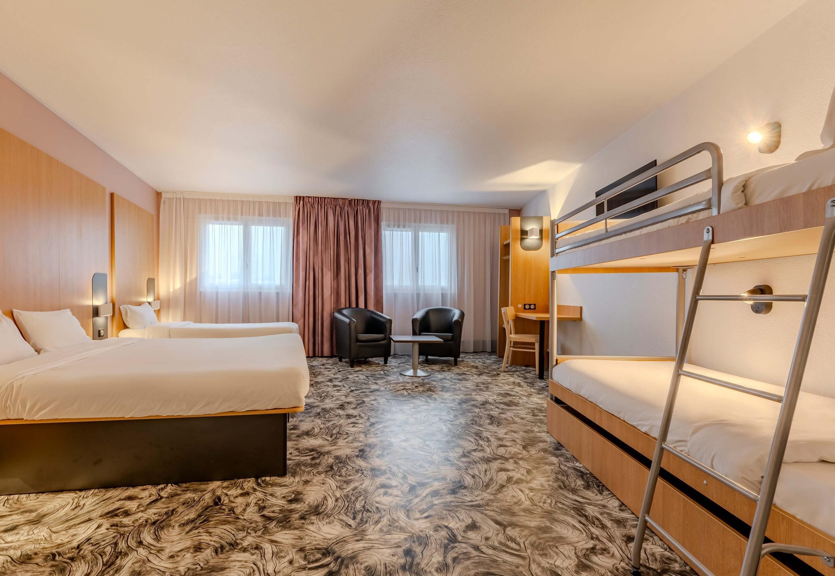 Une chambre d’hôtel avec un lit superposé, un bureau, deux chaises, une petite table et une grande fenêtre avec des rideaux.