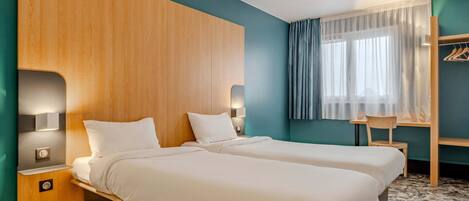 A hotel room with two beds, a wooden headboard, a desk with a chair, and a window with curtains.
