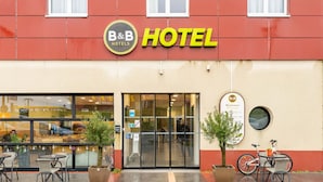 A B&B hotel with a red facade, a glass entrance, outdoor seating, and a bicycle parked outside. - B&B HOTEL Maubeuge Louvroil (Louvroil)