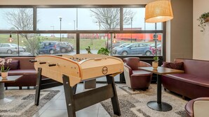 A room with a foosball table, a sofa, a table with a lamp, and a view of parked cars outside. - B&B HOTEL Maubeuge Louvroil (Louvroil)