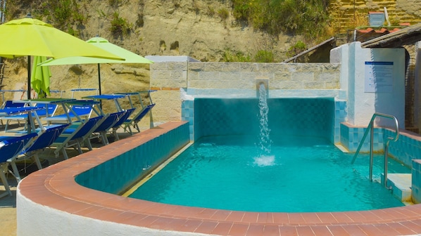 A swimming pool with a water feature, surrounded by blue lounge chairs and a green umbrella. - Hotel Regina del Mare (Barano d'Ischia)