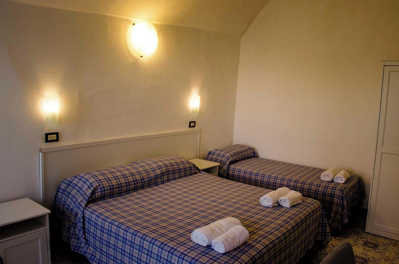 A room with two beds, each with a headboard, a bedside table, and a wall-mounted light fixture.