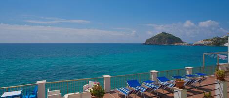 A terrace with blue chairs and tables overlooking a turquoise sea, with a small hill in the background.