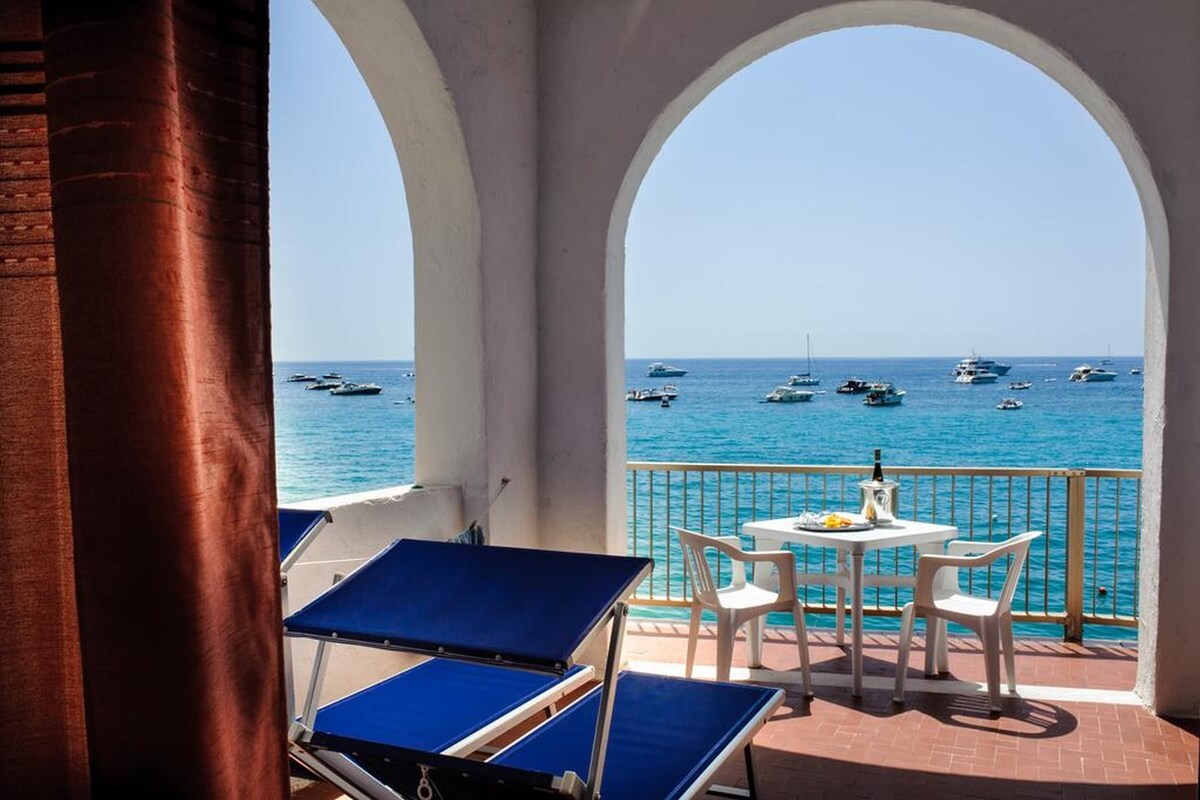 Un balcón con vista al mar, con una mesa, sillas y una tumbona azul.