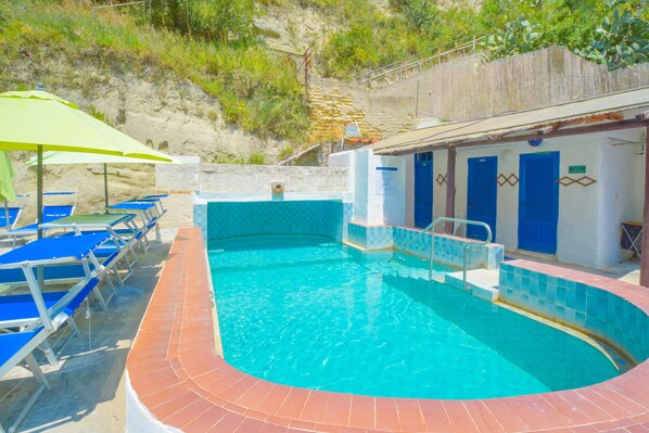 A swimming pool with blue chairs and a green umbrella, surrounded by a stone wall and a building with blue doors.