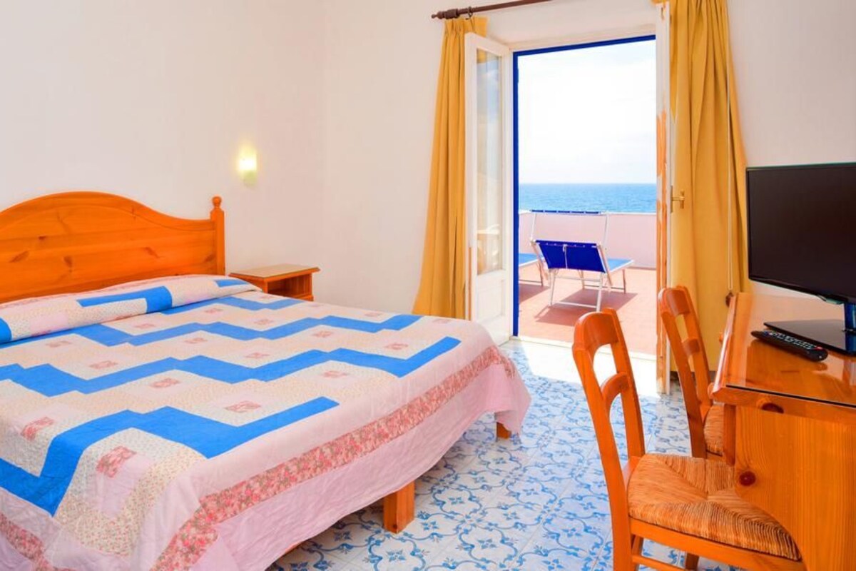 Een slaapkamer met een bed, een televisie, een bureau en een stoel. Er is een balkon met uitzicht op zee.