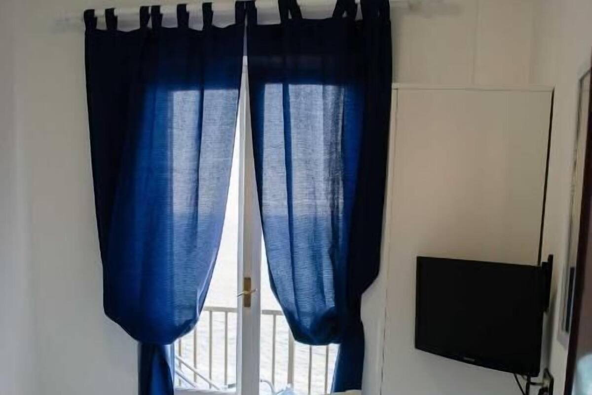 Une pièce comprenant un lit, un bureau, une chaise, un téléviseur et une fenêtre avec des rideaux.