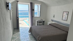 Een slaapkamer met een bed, een televisie, een klein tafeltje en een raam met uitzicht op zee.