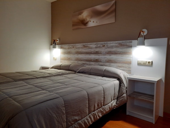 A neatly made bed with a grey quilt, two bedside tables with lamps, and a wall-mounted power outlet. - Hotel Muradana (Muros)