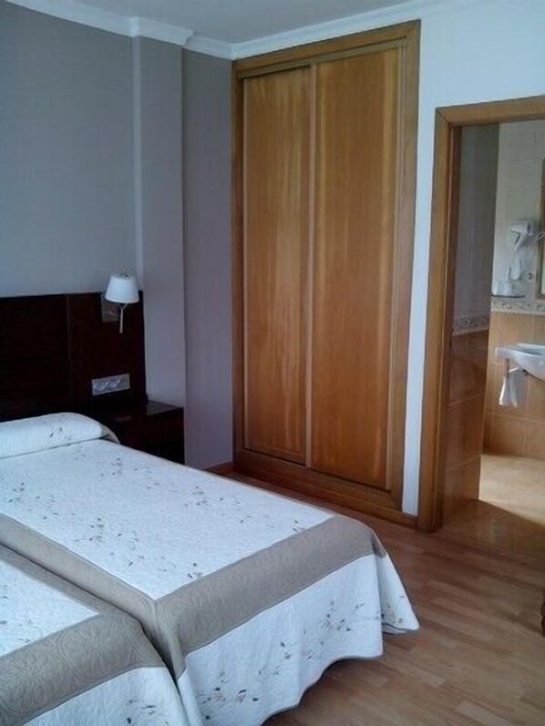 Habitación de hotel con dos camas, un armario de madera, una mesita de noche con lámpara y un baño visible a través de una puerta abierta.