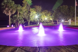 Vista nocturna de una fuente iluminada de morado, rodeada de árboles y un edificio al fondo.
