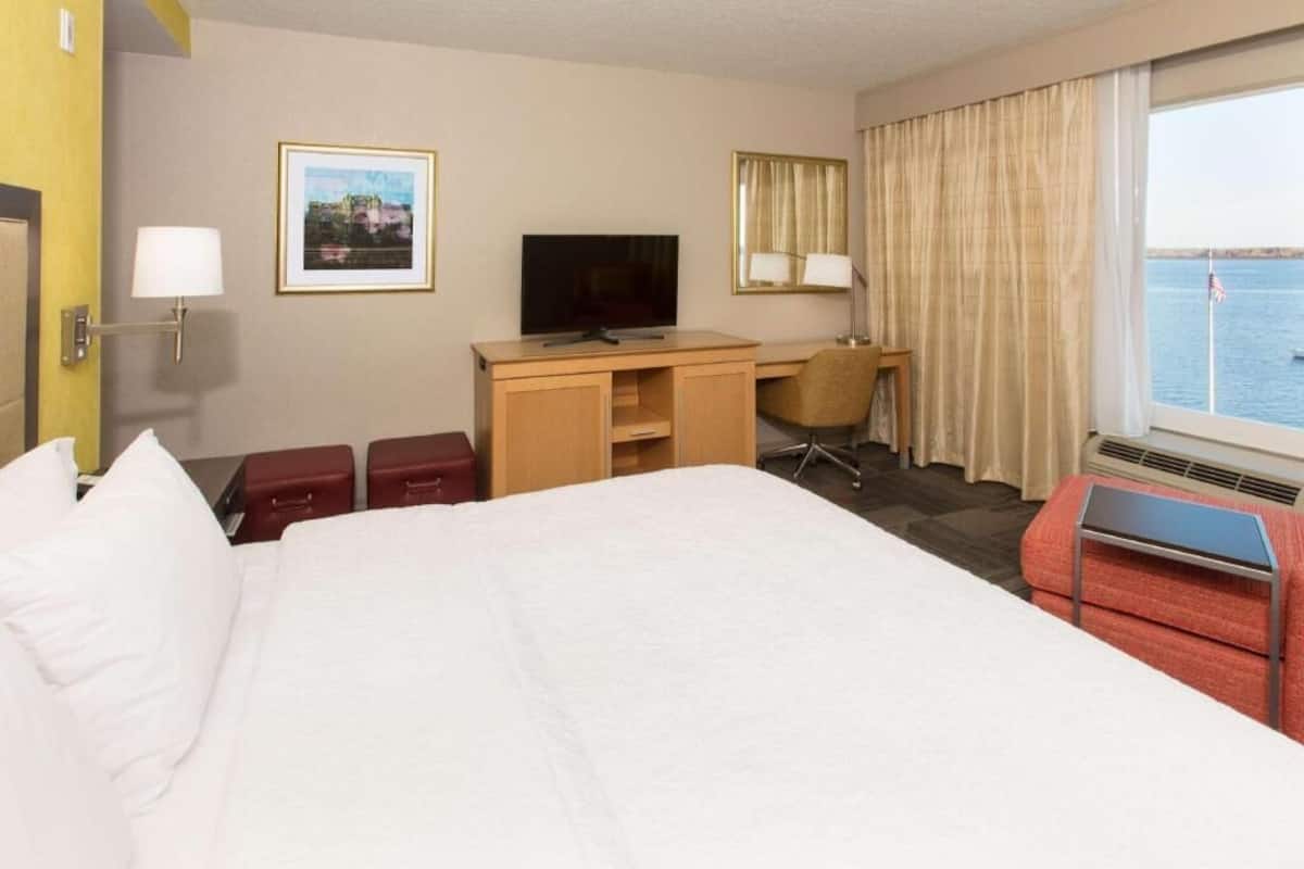 Habitación de hotel con cama, escritorio, silla, televisor y ventana con vista al agua.